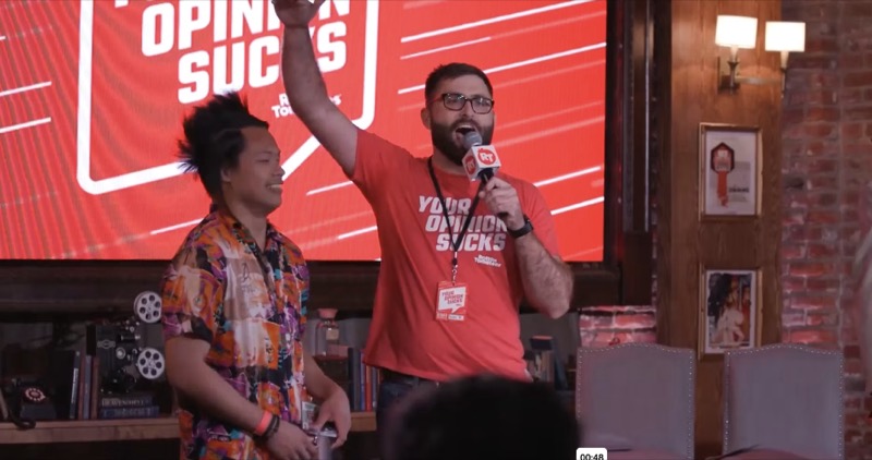 Scott Rowan with a microphone, arm raised, warming up the crowd at Rotten Tomatoes’ Your Opinion Sucks